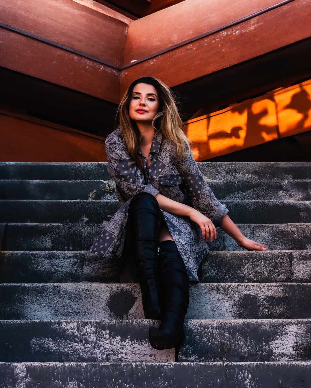 Fashion portrait of a woman in a patterned shirt dress and black thigh-high boots sitting on concrete outdoor stairs bathed in warm sunset lighting.