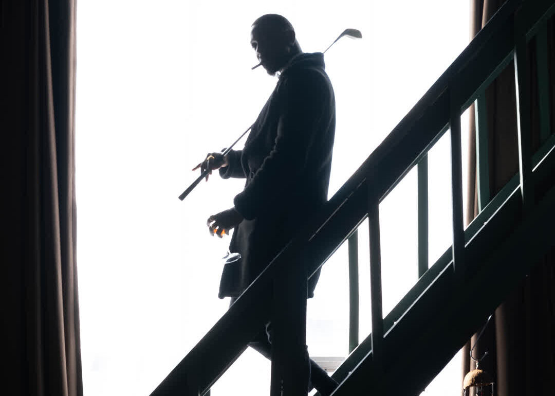 Cinematic silhouette of a man walking down indoor stairs, holding a golf club and a glass while smoking.