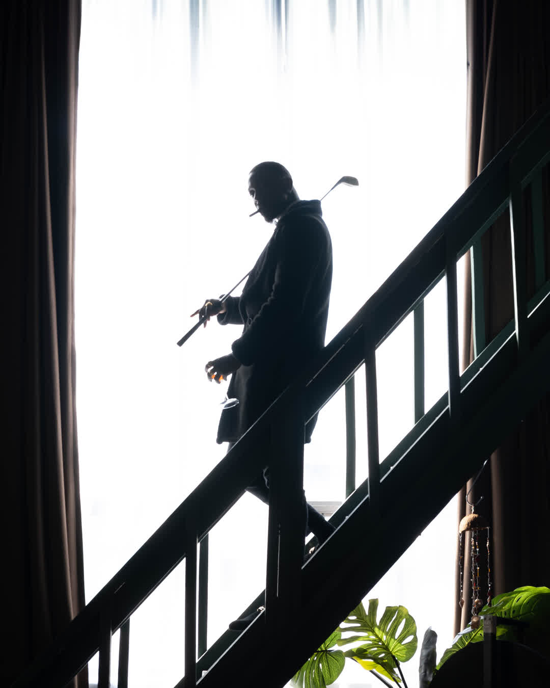 Cinematic silhouette of a man walking down indoor stairs, holding a golf club and a glass while smoking.