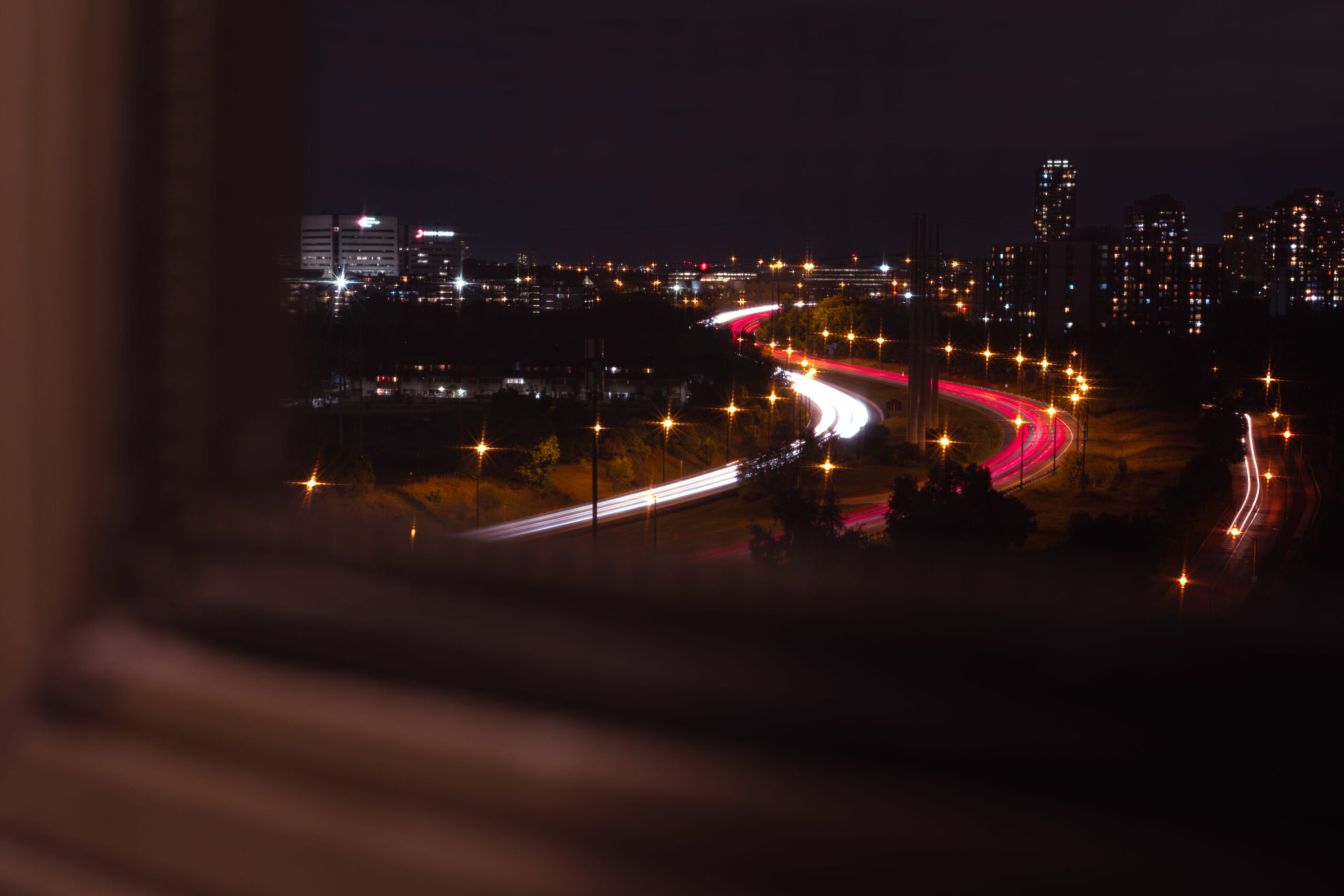 Long exposure night photography of a winding highway with red and white light trails leading into a glowing city skyline.