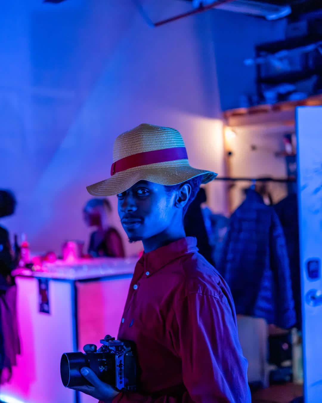 Portrait of a young Black man wearing a straw hat and red shirt holding a mirrorless camera, illuminated by striking blue and purple neon lights at an event.