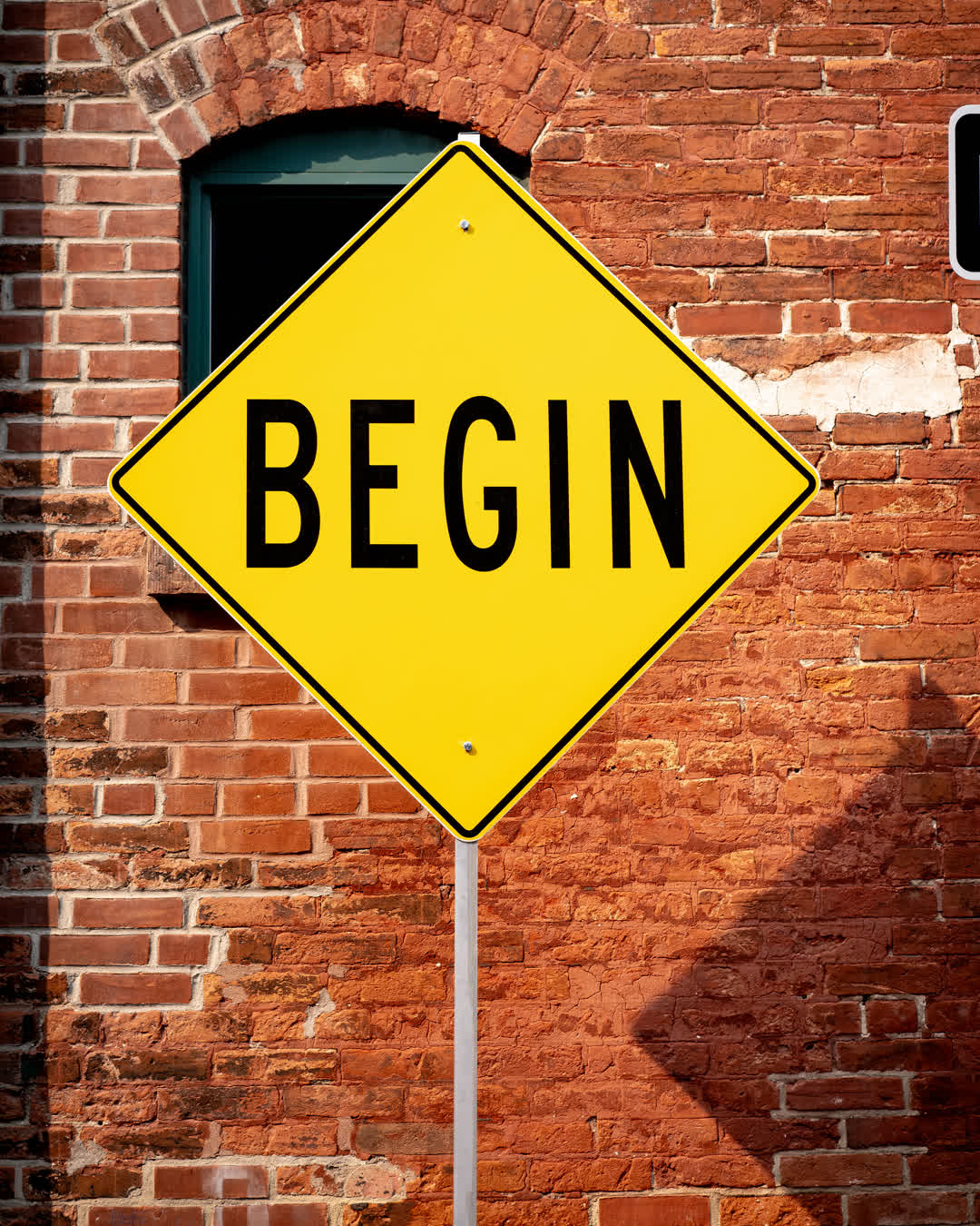 A yellow diamond-shaped street sign reading the word BEGIN in bold black letters, standing in front of an old red brick building.