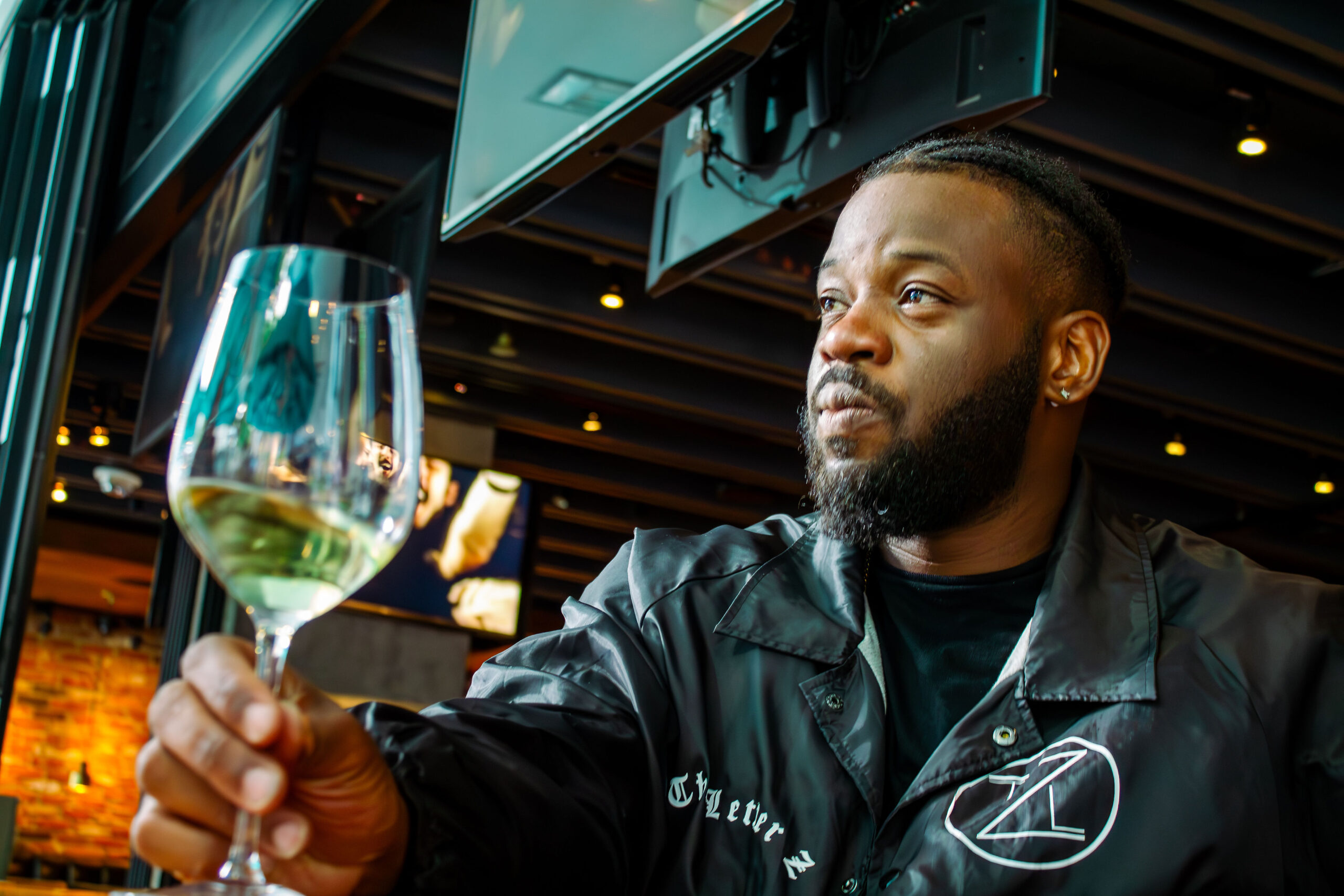A man with a beard sitting at a modern indoor-outdoor bar, looking contemplatively off-camera while holding a glass of white wine in the foreground.