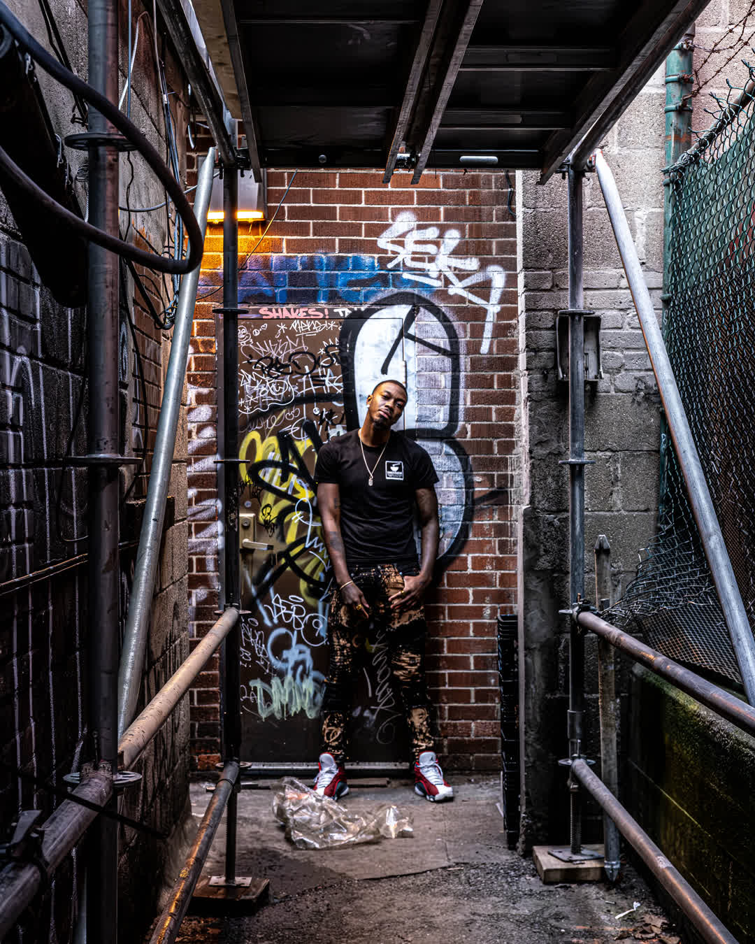 Full body portrait of a confident Black man wearing streetwear and red sneakers, standing in a grungy, graffiti-covered urban alleyway framed by metal scaffolding.