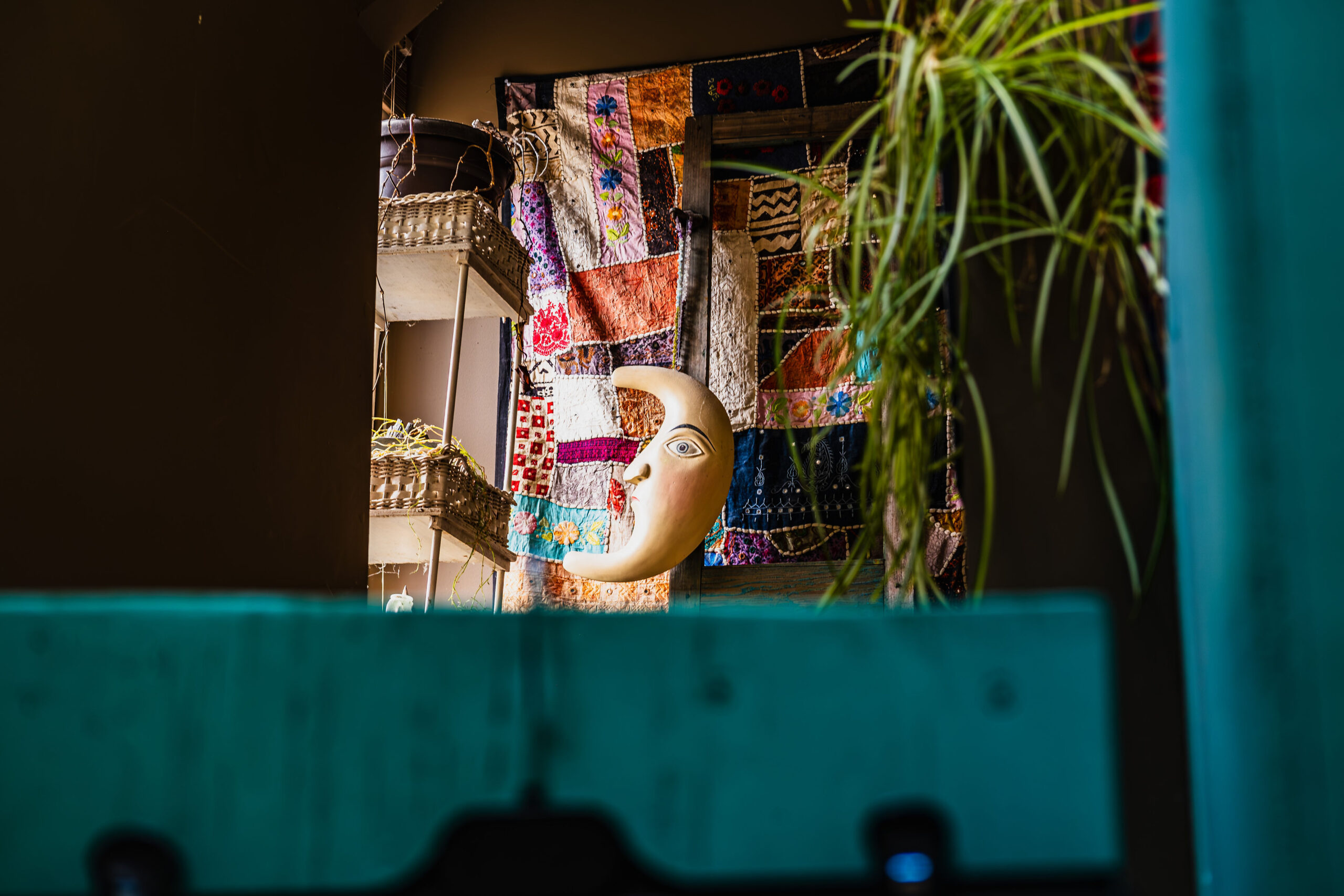 Crescent moon face sculpture hanging in a window against a colorful patchwork quilt, framed by teal foreground elements.