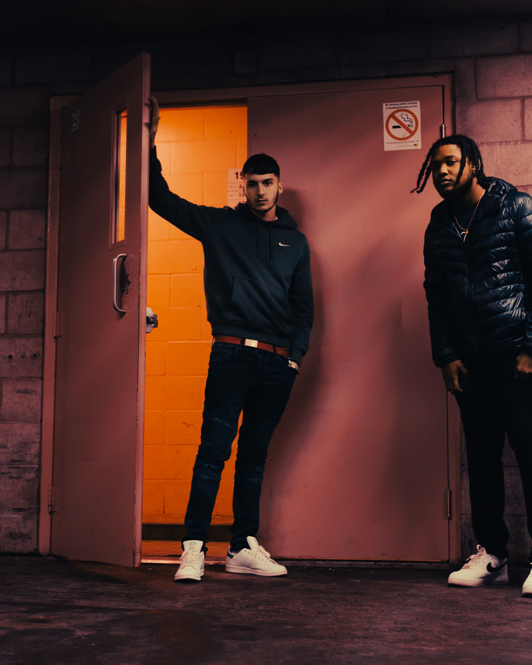 Two young men in streetwear standing confidently by a partially open pinkish-red industrial door with warm interior lighting.