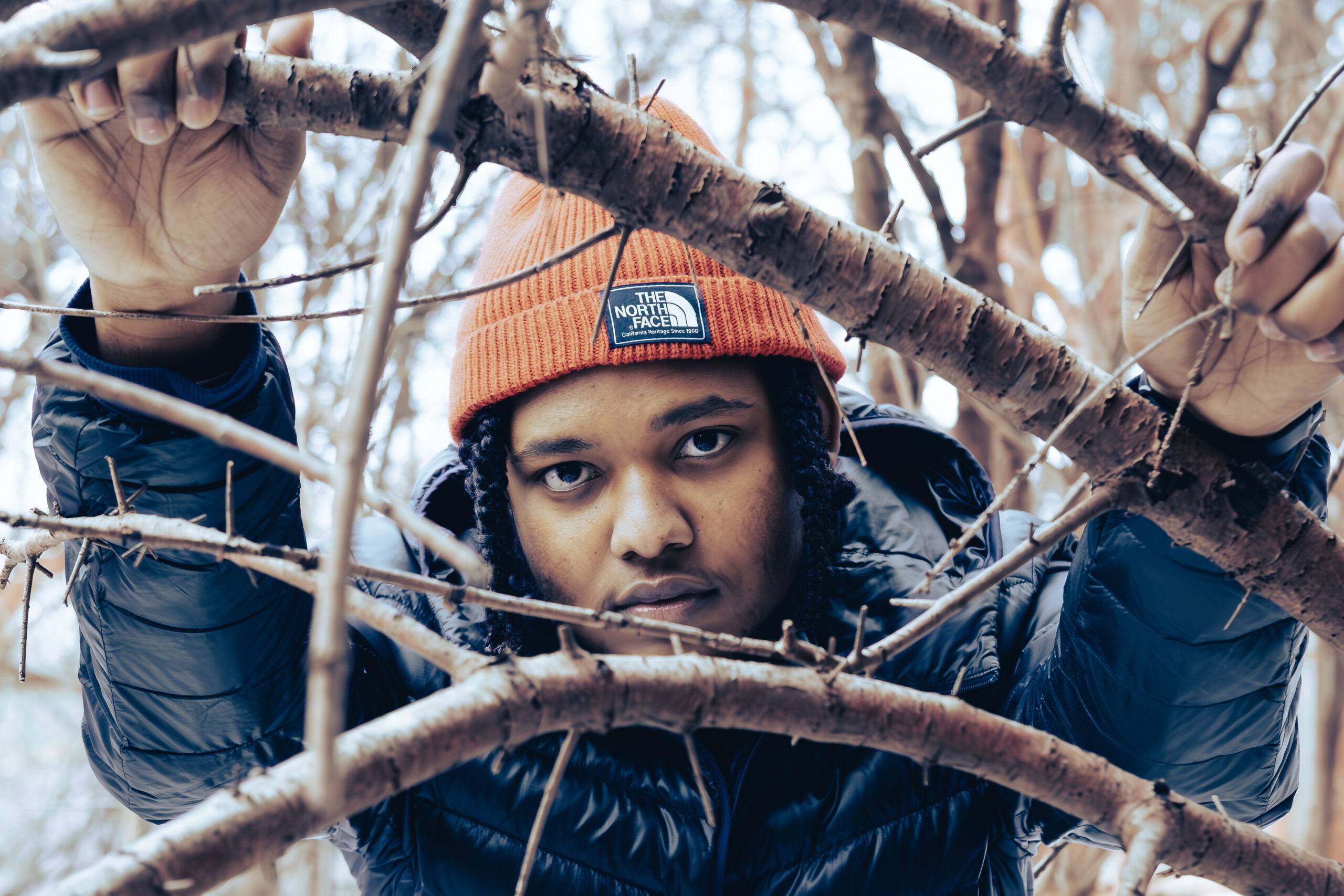Close-up portrait of a person wearing an orange North Face beanie and dark jacket, looking intensely into the lens through a natural frame of thorny branches.