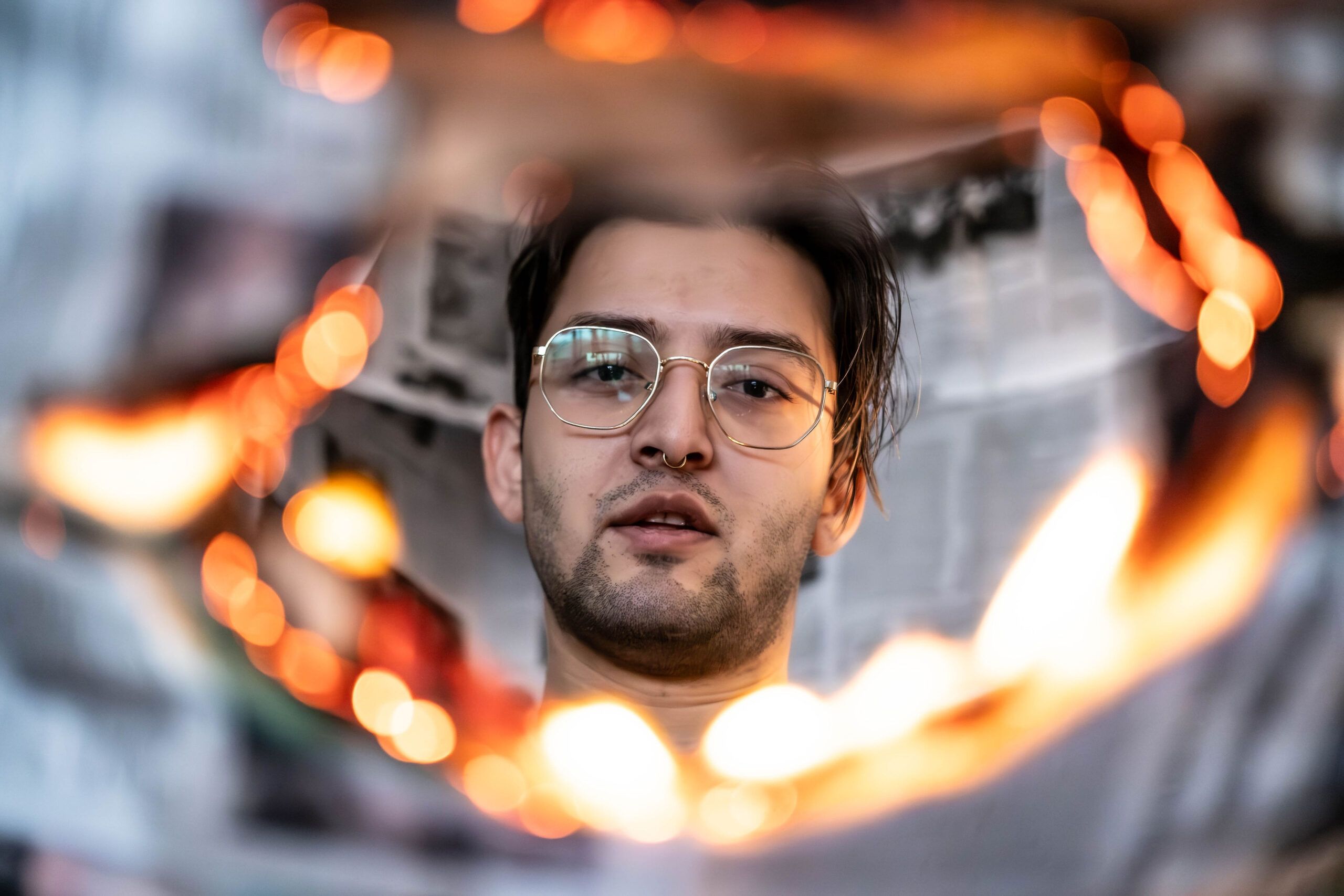 Portrait of a young man with glasses and a septum piercing, looking calmly through a glowing, out-of-focus ring of fire or warm light.