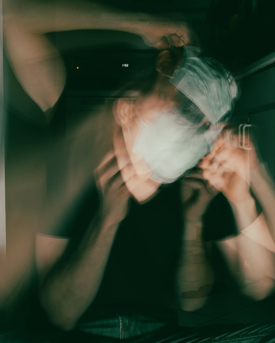 Abstract, blurry, long-exposure photograph of a person adjusting a light blue medical mask, characterized by a greenish, cinematic tint and motion blur.