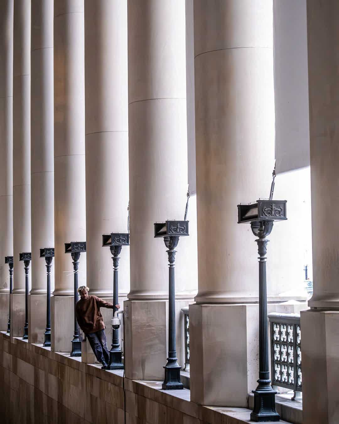 A person in a brown sweater leaning against the base of massive stone pillars in an open-air walkway lined with black, vintage lampposts