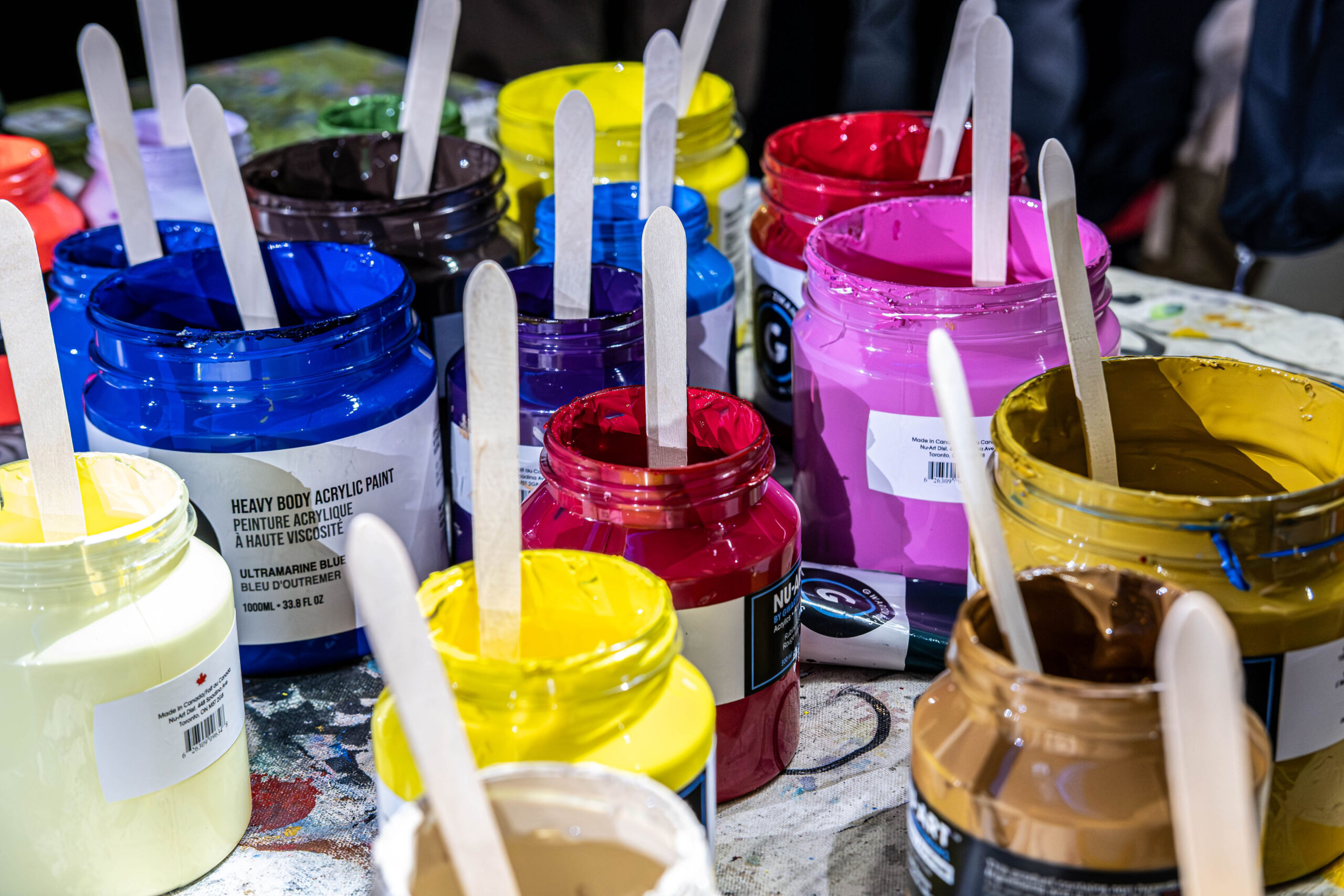Several open jars of bright, heavy body acrylic paint in shades of blue, pink, yellow, and red with wooden stir sticks resting inside them.