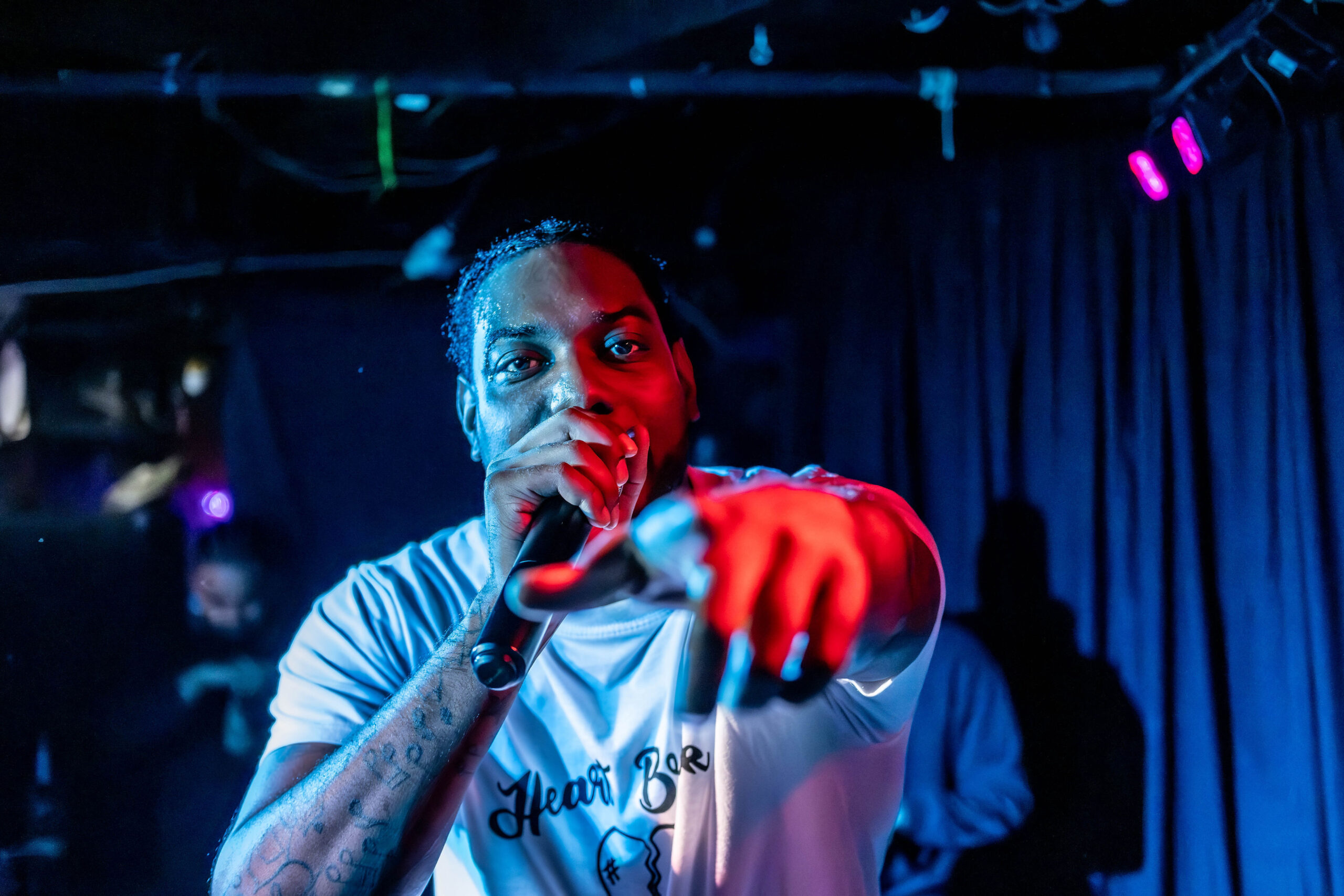 Live performance photograph of a musician holding a microphone and pointing directly at the camera, illuminated by striking red and blue stage lighting in a dark venue.